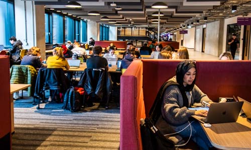 Image shows wide shot of students working in one of the open plan study spaces. In the front of the shot, there are students sat in a red study booth working on a project together. Behind them, more students are working collaboratively.