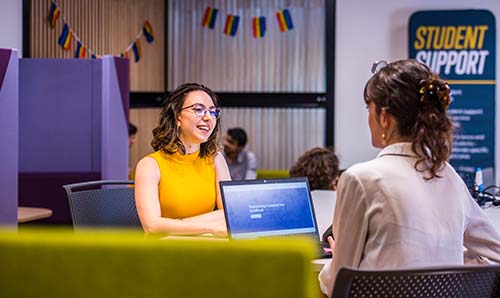 A student is sat at a desk talking with a member of staff from the Student Support Hub about the help available.