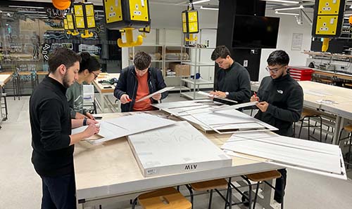 A group of four students are working on a foam board aeroplane wing in our Makerspace. They are working around wooden benches and build the foam wing of a plane for a project.